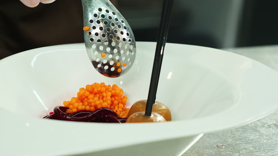 Chef places caviar on dessert plate using slotted spoon closeup. Man places all ingredients on plate creating high-level presentation of dish