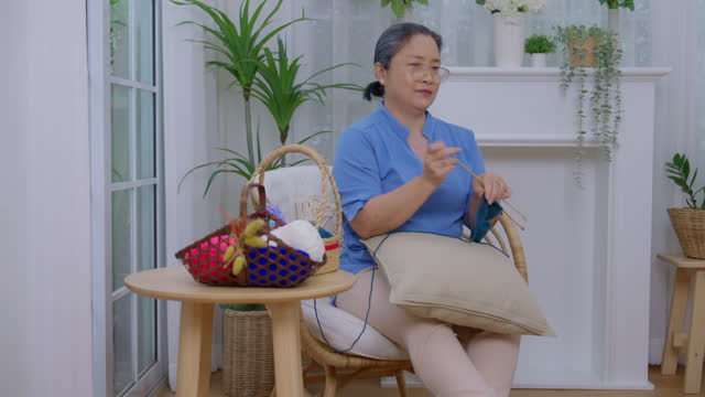 Smiling Asian elderly woman wearing glasses sits on rattan chairs and knits for leisure and retirement hobbies inside the home
