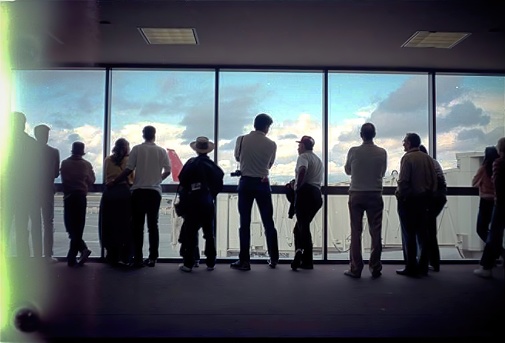 This photograph depicts a group of people standing on a balcony, gazing out towards the airport runway, possibly in anticipation of an arriving flight.
