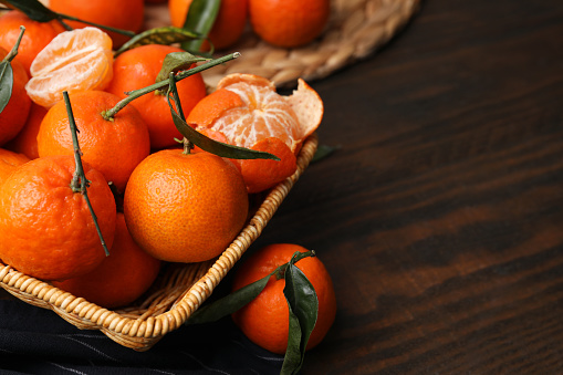 Fresh ripe tangerines with green leaves in wicker basket on wooden table, closeup. Space for text