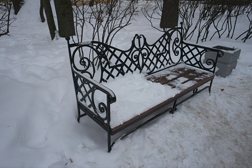 Beautiful wrought iron bench in a winter city park among the trees, a curly back with an intricate pattern, a wooden seat covered with snow, a trash can nearby, seasonal landscape, parks and nature, vintage garden furniture, cloudy day, winter.