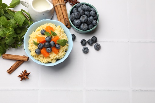 Tasty millet porridge with blueberries, pumpkin and mint in bowl on white tiled table, flat lay. Space for text