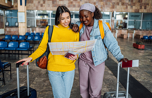 Young cheerful multi-ethnic female friends with suitcases and passports looking for directions on a map at the train station