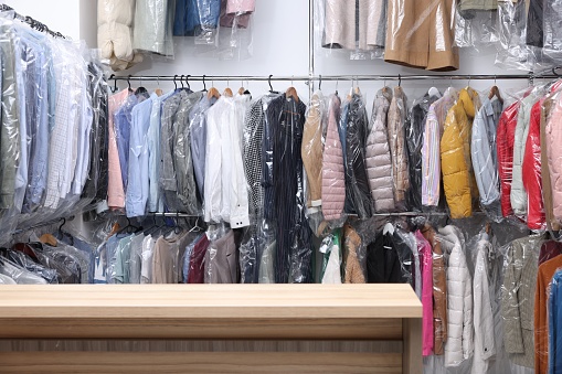 Dry-cleaning service. Hangers with different clothes in plastic bags on racks and wooden counter indoors