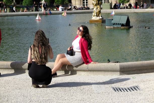 Paris, France - Oct. 4, 2023:  A young woman poses for a photo on a sunny afternoon in the Jardin du Luxembourg, sitting on the edge of the pond in front of the Palais du Luxembourg, on the Left Bank of the Seine River.