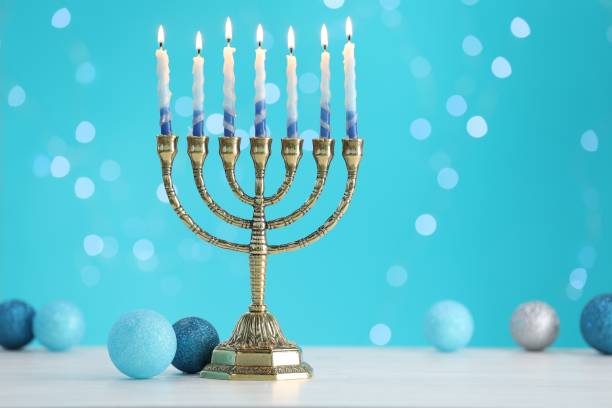 istock Hanukkah celebration. Menorah with burning candles and baubles on table against light blue background with blurred lights, space for text 1890507199