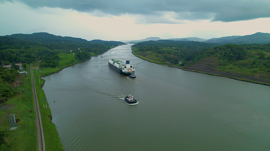A large cargo ship in transit through the man-made waterway of Panama Canal. An escorted freighter travels along a strategic water shortcut that connects two oceans. Remarkable engineering project.