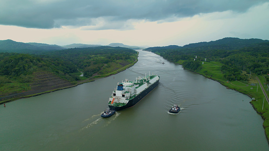Escorted cargo ship travels between two oceans through Panama Canal waterway. Strategic man-made water shortcut divides Central and South America and enables shortening of routes for maritime traffic.