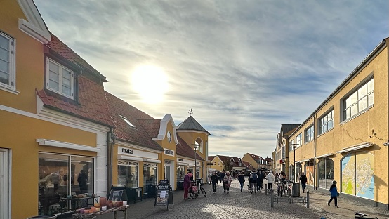 Skagen, Denmark - October 18, 2023: In late October, the town center buzzes with life under a crisp, sunny afternoon sky, the low-angled sun casting long shadows across the cobbled streets. Locals and visitors alike mingle among the quaint cafes and colorful shops, enjoying the cool air and the town's vibrant, yet laid-back atmosphere, accentuated by the rich autumnal colors.