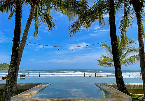Dreamlike view of an oceanfront infinity pool in the shade of exotic palm trees. Stunning location of a resort with swimming pool next to ocean beach. Relaxing holiday retreat in tropical paradise.