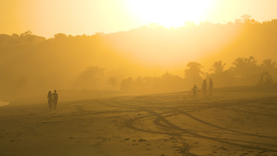 Silhouettes of unrecognizable people walking on misty sandy beach in golden sunlight. Young couple and family having fun while strolling at Playa Venao beach. Leisure activities during summer holiday.
