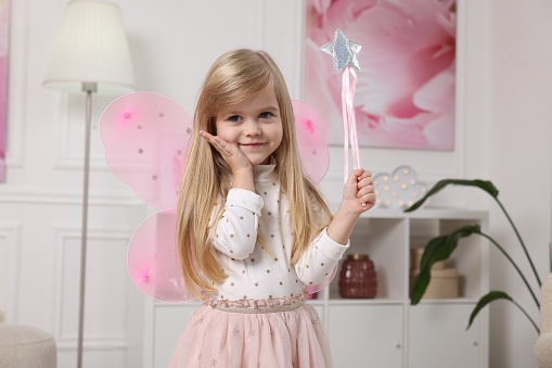 Cute little girl in fairy costume with pink wings and magic wand at home