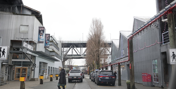 Vancouver, Canada - December 21, 2023: Pedestrians walk among the Granville Island buildings housing cultural institutions, industries, restaurants and retailers. The Granville Street Bridge carries Highway 99 in the background. Winter morning with overcast skies.
