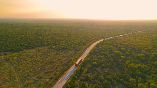 AERIAL: Red container truck driving along Adriatic landscape in golden sunlight. Big delivery truck transporting goods through beautiful countryside with lush green shrubbery in early morning light.