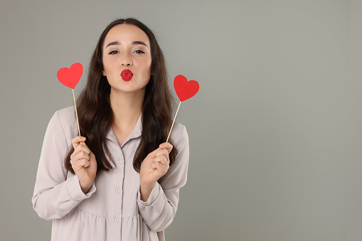 Beautiful young woman with paper hearts sending kiss on grey background, space for text