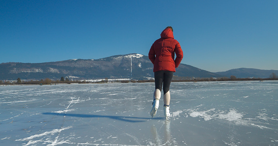 Back view of a sporty woman ice skating across naturally frozen lake under a snowy mountain on a sunny day. A wonderful outdoor sports activity for cold winter days in the beautiful wintry countryside