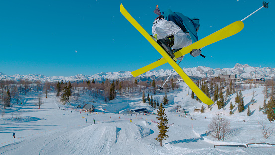 CLOSE UP: Stunning view of freestyle skier doing grab trick while jumping kicker. Young extreme athlete flying high above snow park and ski area. Adrenaline activity on sunny winter day in ski area.