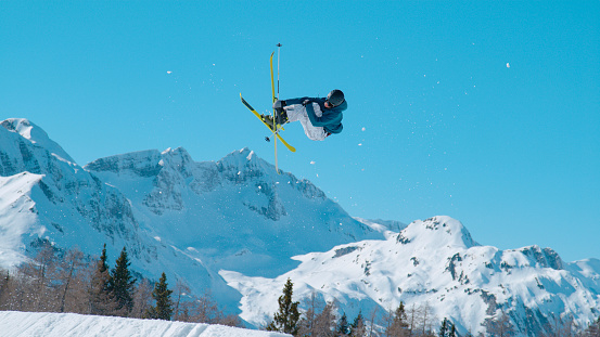LOW ANGLE VIEW: Male freestyle skier flying above snow-covered mountaintops. Young extreme athlete jumping big air kicker at snow park in ski area. Adrenaline activity on sunny winter day in ski area.