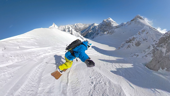 Extreme snowboarding of a man riding fresh snow down a steep mountain slope. Magnificent winter day for riding snowboard and spraying clouds of powder snow in untouched snowy mountain environment.