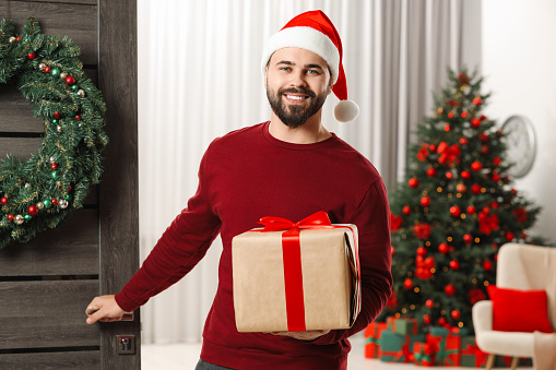 Young man in Santa hat with Christmas gift box indoors. Sending present by mail