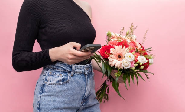 junge kaukasische frau in denim hält telefon mit strauß frischer blumen rosen, gerbera, chrysantheme auf rosa hintergrund. online-bestellung und blumenlieferkonzept. horizontale ebene - rose flower shop variation color image stock-fotos und bilder