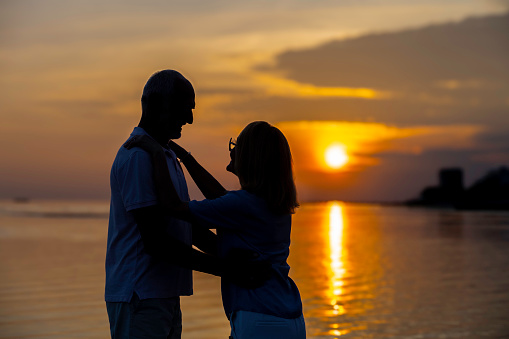 Romantic senior couple silhouettes hugging and looking at each other at coastline at sunset, side view medium shot