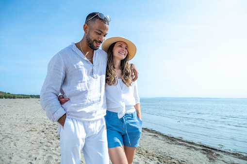 Happy beautiful young multiracial boyfriend and girlfriend walking on beach at sunny day, medium shot