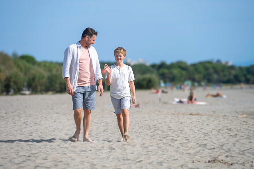Smiling dad and son walking on sand beach barefoot and talking, wide shot front view