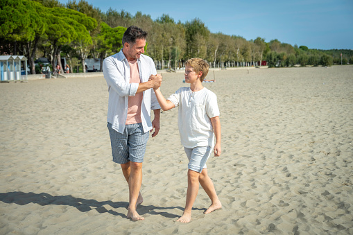Caucasian father and son walking on beach barefoot, chatting and shaking hands, full length shot