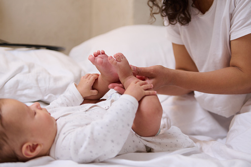 Close-up of a young woman mother massaging feet and toes of her newborn baby, stroking and kissing legs of her baby boy, enjoying happy time together. Family. Motherhood. Baby care and infancy concept