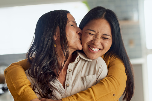 An Asian mother is hugging her adult daughter tightly and kissing her on the cheek. They are both smiling and look happy.