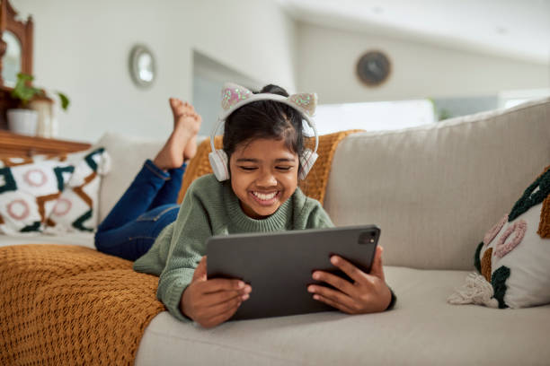 Young Girl Lying on Couch Watching Videos on Tablet An adorable young girl is lying on the couch, engrossed in watching videos on her tablet. She has cat-ear headphones and a big smile on her face. free girl download stock pictures, royalty-free photos & images