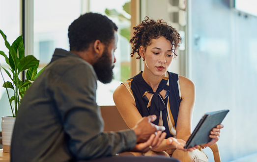 A man and a woman are sitting on a couch and looking at a tablet. They are both dressed in business casual attire. The man has a beard and the woman has curly hair. The background is blurry and there are plants visible.