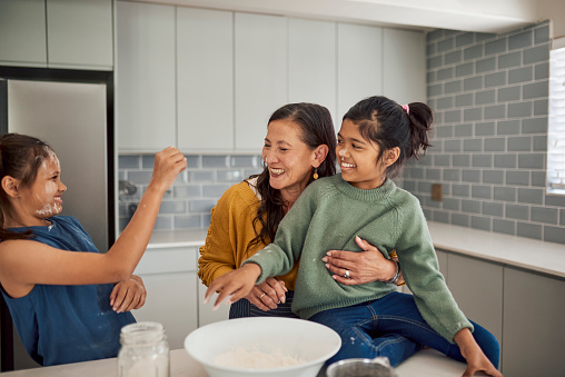 An elated grandmother and her two granddaughters are covered in flour while having fun baking together in a modern kitchen.