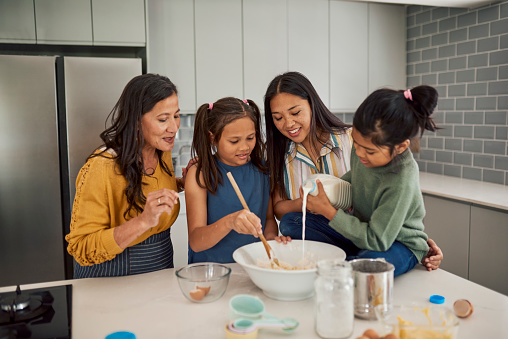 A grandmother, mother, and two daughters bake together in the kitchen. They are smiling and laughing while they mix ingredients and pour milk into a bowl.
