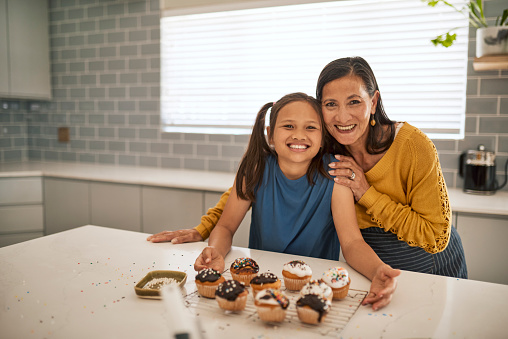 An grandmother and her granddaughter are baking cupcakes together in the kitchen. They are both smiling and look happy.