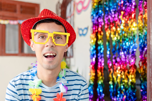 Young Brazilian man in festive Carnival attire, wearing a hat and playful glasses, smiles at the camera. Vibrant and ready for the celebration.