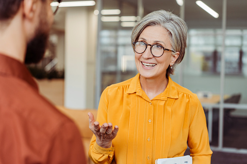 Portrait of smiling confident senior woman wearing eyeglasses communication with colleague at workplace. Business people talking, cooperation, planning startup working in office. Meeting, teamwork