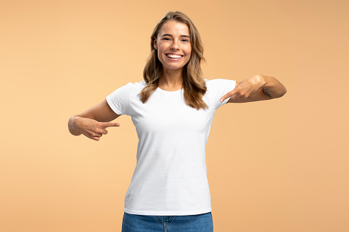 Portrait of smiling attractive woman wearing white t shirt pointing fingers mockup, copy space, looking at camera isolated on beige background. Advertisement concept