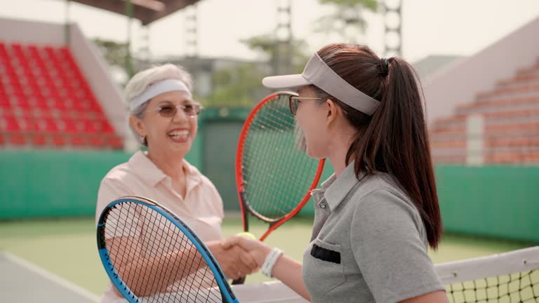 Two Asian senior women tennis players, shaking hands after playing tennis game in stadium. Smiling athletes team standing and using hand gesture and handshake over tennis net. Happy senior friends shaking hands after tennis match in morning.