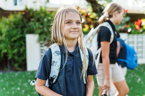 A young girl of Caucasian ethnicity carrying a backpack smiles as she stands outside with fellow students after school.