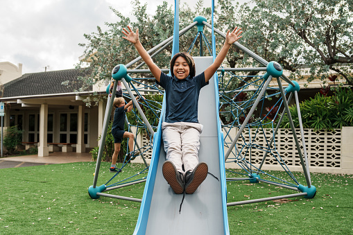 A young schoolboy of Asian ethnicity smiles and excitedly puts his hands in the air as he slides down a play structure slide outside on a cloudy day.