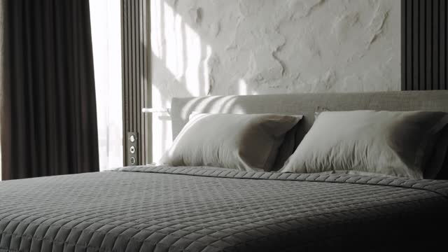 Close-up of bed with white pillows and of gray blanket, sun rays trough the window in large bedroom, bedding, slow motion.