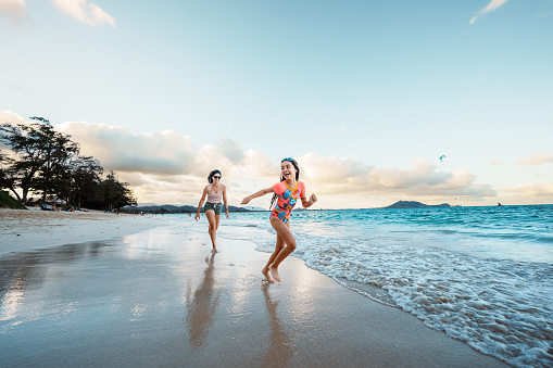 An adorable elementary school aged girl wearing a swimsuit smiles as she and her mother run down the beach into the ocean together in Hawaii.