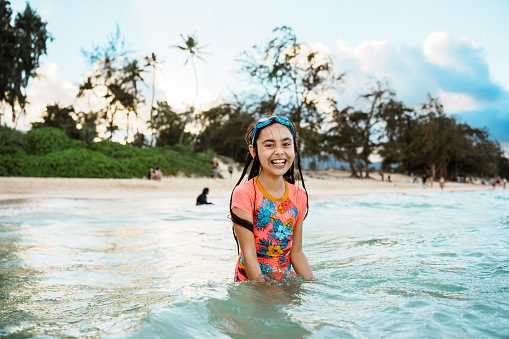 A smiling elementary school age girl looks directly at the camera as she stands in the ocean while enjoying the weekend outside.