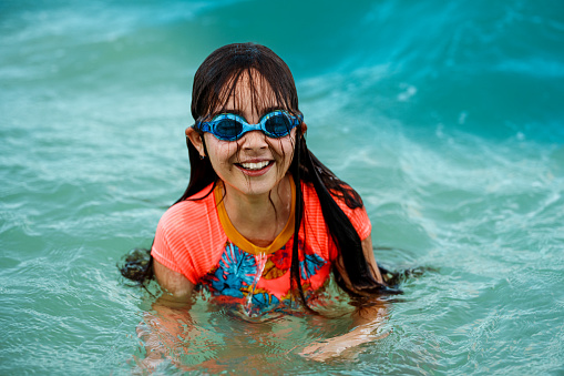 An elementary school age girl of Caucasian descent wearing a swimsuit and goggles, swims in the ocean water and smiles directly to the camera.