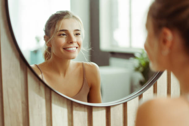 smiling, confident woman with make up wearing tank top, looking in mirror, standing in bathroom - women body beauty candid imagens e fotografias de stock