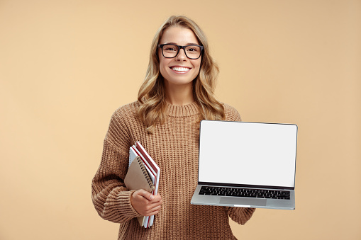 Portrait of smiling beautiful woman wearing glasses, holding laptop, showing mockup, looking at camera standing isolated on beige background. Online education concept, technology