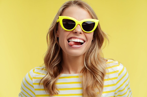 Portrait of beautiful smiling woman wearing stylish yellow sunglasses showing tongue out, fooling around, standing isolated on yellow background. Advertisement concept