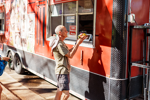 A senior man of Caucasian descent stands at the window of a food truck, receiving a fresh açaí bowl on a beautiful sunny weekend day in Hawaii.